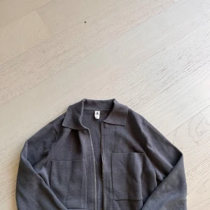 Grå stickad overshirt från Åhléns - Säljer en stilren grå stickad overshirt från Åhléns med dragkedja framtill och två stora fickor. Jackan har klassisk krage och är långärmad, perfekt för lager-på-lager. Enkel och clean look som funkar till mycket.