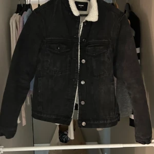 Svart teddyjeansjacka från BikBok XS - Svart jeansjacka från BikBok i storlek XS med vit teddyfoder på insidan och kragen. Klassisk modell med bröstfickor, silverfärgade knappar och långärmad passform. Perfekt för dig som vill ha en cool och mysig jacka med streetkänsla.
