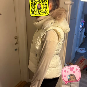 Vit dunväst från Moncler med pälskrage - Snygg vit dunväst från Moncler med stor fluffig pälskrage på huvan. Västen har dragkedja framtill, två fickor med dragkedja och en markerad midja. Perfekt för lager-på-lager och ger en lyxig vibe till din outfit.