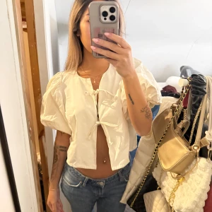 Vit blus med puffärm och knyt - Säljer en superfin vit blus med korta puffärmar och två knyt framtill. Blusen är croppad och har en luftig passform som ger en cool och trendig vibe. Perfekt till jeans eller kjol för en snygg look.