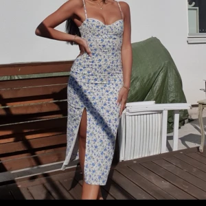 Blommig långklänning med slits - Supersöt långklänning med smala axelband och figurnära passform. Klänningen är vit med blått blommönster och har en snygg slits framtill. Söt sweetheart-ringning och lätt rynkade detaljer vid bysten. Perfekt för sommarens alla tillfällen!