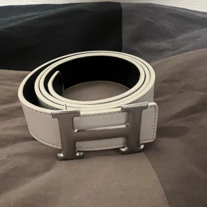 Vitt skärp med H-spänne - Säljer ett stilrent vitt skärp med ikoniskt H-spänne i silverfärgad metall. Bältet är gjort i slätt skinn och har en bred design som verkligen sticker ut. Perfekt accessoar för att lyfta vilken outfit som helst. Storlek 105cm, använd bara en gång. 