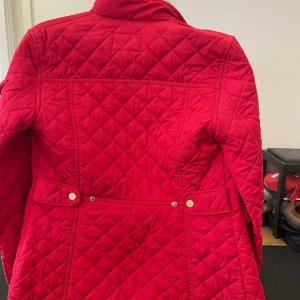 Röd quiltad jacka med fickor - Säljer en snygg röd quiltad jacka med fyra fickor framtill och guldiga knappar. Jackan har hög krage med spänne, dragkedja och tryckknappar. Perfekt för dig som vill sticka ut med färg och stil under kyligare dagar.
