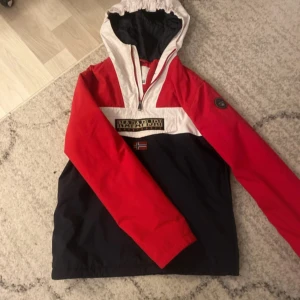 Röd, vit och marinblå vindjacka Napapijri - Säljer en snygg vindjacka från Napapijri i rött, vitt och marinblått. Jackan har huva, halv dragkedja och patch med logga på ärmen. Klassisk streetstil med norsk flagga framtill och stor logotyp. Perfekt för blåsiga dagar.