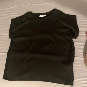 Svart blus från VILA, storlek 44 - En stilren svart blus från VILA i storlek 44. Blusen har en rak och loose passform med korta ärmar och rund halsringning. Materialet är mjukt och har en lätt glansig finish som ger en snygg look. Perfekt att styla med jeans eller kjol.
