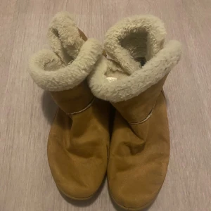 Uggs - Mysiga beiga boots med mjukt vitt foder i fårskinn och ovandel i mocka. Skorna har rund tå och platt sula, perfekta för kalla dagar. Klassisk design med uppvikt kant och sömdetaljer. Säljs i par.