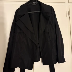 Svart trenchcoat från Boohoo - Svart trenchcoat från Boohoo med bred krage, långa ärmar och bälte med metallöljetter i midjan. Jackan har en loose passform och är tillverkad i ett lätt syntetmaterial. Perfekt för dig som gillar stilrena och klassiska vibes.