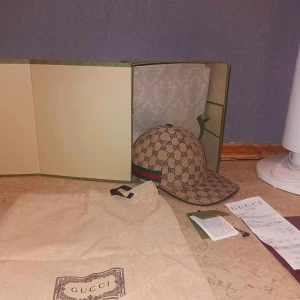 Gucci keps med GG-mönster och röd-grön rand - Exklusiv keps från Gucci i beige med klassiskt GG-monogram över hela kepsen och en ikonisk röd-grön rand på sidan. Kepsen är tillverkad i bomullscanvas med svarta detaljer på insidan och justerbar rem bak. Kommer med originalpåse, box och kvitto.