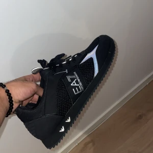 Svarta sneakers från Emporio Armani EA7 - Snygga svarta sneakers från Emporio Armani EA7 med silvriga detaljer och reflekterande inslag. Ovandelen är i mesh och syntet, med tydlig logga på sidan. Skorna har snörning och en räfflad platt sula för extra grepp. Perfekta för dig som vill ha en sportig och stilren look.