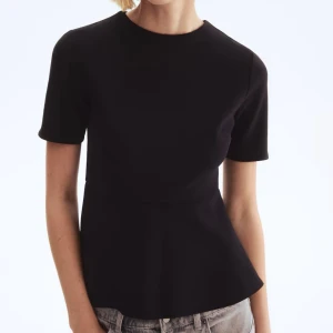 Svart peplum topp med kort ärm - Snygg svart topp med peplumskärning och rund hals. Toppen har korta ärmar och en figurnära passform som ger en stilren siluett. Perfekt att matcha med jeans eller kjol för en trendig look.