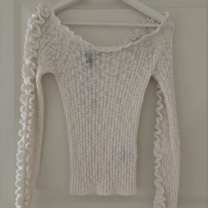 Vit ribbad stickad tröja - Supersöt vit ribbad stickad tröja från Lindex. Tröjan har långärmade ärmar med volangdetaljer längs axlar och ärmar samt en bred, något båtringad hals. Aldrig använd. 