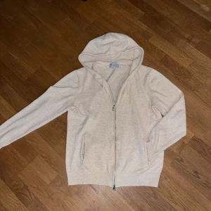 Beige Kashmir tröja  - Beige kashmir tröja med dragkedja framtill och huva med snörning. Tröjan har långa ärmar, ribbade muddar och två fickor framtill. Perfekt för en chill och avslappnad stil.