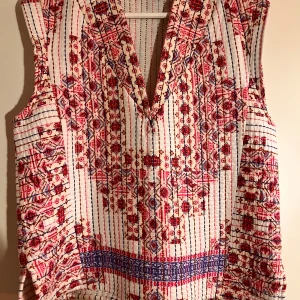 Mönstrad ärmlös blus från Zara - Säljer en ärmlös blus från Zara med v-ringning och färgstarkt mönster i rött, blått och vitt. Blusen har broderade detaljer och ett bohemiskt mönster med inslag av ränder och små blommor. Tillverkad i mjuk bomull, perfekt för varma dagar.