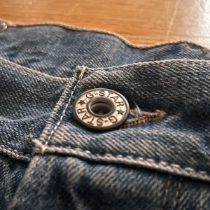 G-Star RAW blå jeans med unika detaljer - Säljer ett par blå jeans från G-Star RAW med snyggt slitna detaljer och unika bakfickor. Jeansen har klassisk femficksdesign, knappar framtill och tydliga kontrastsömmar. Märkespatch på bakfickan och cool faded effekt på benen. OBS! Benen är uppsydda så skriv angående specifika mått!