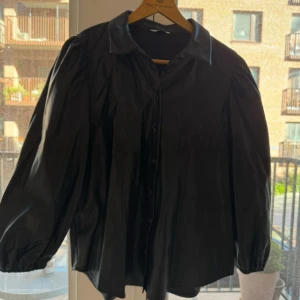 Svart skjorta i skinnimitation från ONLY - Svart skjorta i skinnimitation från ONLY med klassisk krage och knäppning framtill. Skjortan har långa puffärmar med resår vid ärmslut.