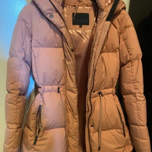 Rosa pufferjacka från Áhkká, strl 34 - Lång, puffig vinterjacka från Áhkká! Den är i mycket bra skick, använd en säsong. De två fickorna utanpå är väldigt stora och så finns det även en ficka inne i jackan på vänster sida. Man kan dra åt i midjan, längs ner och även i luvan. Bandet som man hänger upp jackan med är av (titta på bild 3) och den är lite smutig vid ärmarna annars mycket bra skick!  90% dun. 10% fjäder. Den har kostat 2800kr. Skriv för fler bilder eller vid frågor!!💕
