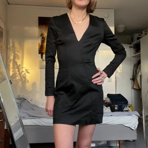 Svart kort klänning med v-ringning - Stilren svart kort klänning med lång ärm och djup v-ringning. Figurnära passform som ger en snygg siluett. Tillverkad i ett slätt, lätt glansigt material som känns lyxigt. Perfekt för dig som gillar minimalistisk och elegant stil.