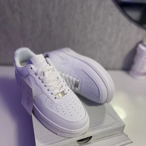 Nike Air Force 1 helvita sneakers - Klassiska Nike Air Force 1 sneakers i helvitt skinn med perforerad tå, snörning och låg profil. Snygg och clean design med Nike-logga på sidan och bak. Perfekta för dig som vill ha en tidlös och fräsch look till vardags.