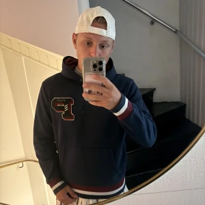 Marinblå hoodie Polo Ralph Lauren M - Säljer en marinblå hoodie från Polo Ralph Lauren i storlek M. Tröjan har en stor broderad bokstav på bröstet, huva och ribbade muddar med röd och vit detalj. Perfekt för dig som gillar klassisk och sportig stil.