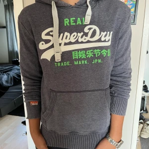 Grå Superdry hoodie - Mörkgrå hoodie från Superdry med vit och grön logga framtill. Tröjan har huva med breda snören, känguruficka och ribbade muddar vid ärmslut och nederkant. Perfekt för chill dagar och streetstyle.