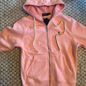 Rosa hoodie från Polo Ralph Lauren - Säljer en ljusrosa hoodie från Polo Ralph Lauren med dragkedja och huva. Tröjan har vita snören, två fickor framtill och broderad logga på bröstet. Perfekt för en chill och avslappnad stil. Det är storlek M men sitter mer som en S