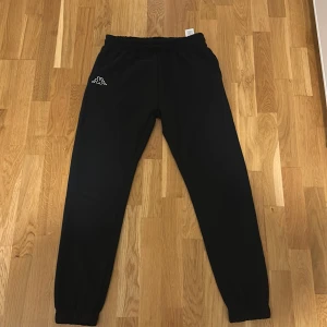 Svarta mjukisbyxor från Kappa - Svarta mjukisbyxor från Kappa med klassisk logga broderad på vänster lår. Byxorna har resår i midjan och vid bensluten samt snörning i midjan. Tillverkade i en mjuk blandning av polyester och bomull för maximal komfort.