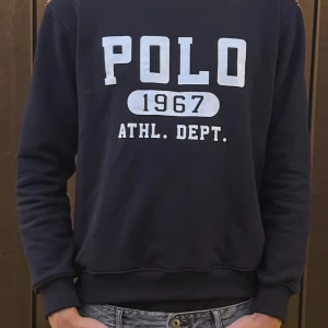 Svart sweatshirt från Polo Ralph Lauren - Säljer en svart sweatshirt från Polo Ralph Lauren med vit tryckt text 'POLO 1967 ATHL. DEPT.' på bröstet. Tröjan har rund halsringning, ribbade muddar och är i mjuk bomull. Perfekt för en avslappnad och sportig stil.