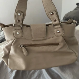 Beige handväska med snyggt spänne - Stilren beige handväska med två rejäla handtag och ett dekorativt silverfärgat spänne framtill. Väskan har mjukt, lätt glansigt material och veckade detaljer som ger en cool look. Perfekt för dig som vill ha en klassisk men trendig accessoar.