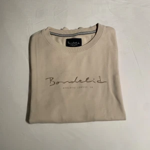 Beige sweatshirt från Bondelid - Snygg beige sweatshirt från Bondelid med broderad logga på bröstet. Tröjan har rund hals, ribbade muddar och är långärmad. Perfekt för dig som gillar stilrena och klassiska plagg med en modern touch.