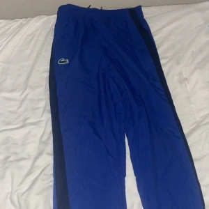 Blå täckbyxor från Lacoste Sport - Säljer ett par blå täckbyxor från Lacoste Sport med klassisk krokodillogga på benet och svarta sidopaneler. Byxorna har elastisk midja med snörning och är gjorda i lätt syntetmaterial, perfekta för sport eller chill. Sköna och stilrena för dig som gillar streetwear.