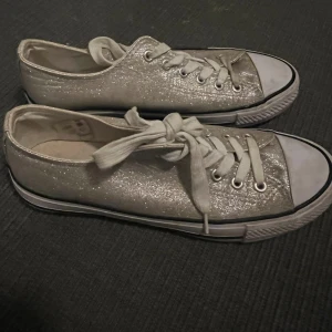 Glittriga silverguldiga sneakers - Säljer ett par snygga sneakers i guld med glittrig finish och vita snören. Skorna har klassisk rund tå och platt sula, perfekta för dig som vill sticka ut lite extra. Passar till både jeans och kjol för en cool look.
