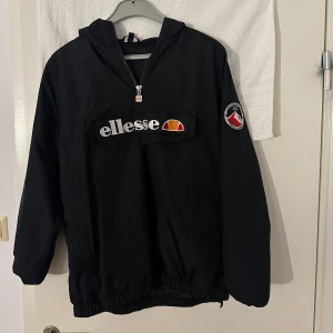 Svart vindjacka från Ellesse - Svart vindjacka från Ellesse med huva och halv dragkedja. Stor logotyp framtill och patch på ena ärmen. Jackan har elastisk nederkant och ärmar, samt en stor ficka på magen. Perfekt för dig som gillar sportig och clean stil.