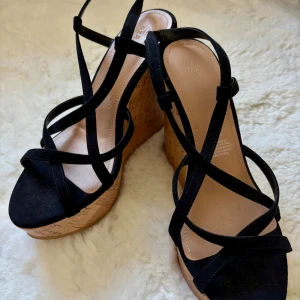 Svarta kilklackssandaler från H&M strl 38 - Snygga svarta sandaler från H&M med högre kilklackar i korklook. Skorna har smala remmar i mockaimitation och öppen tå, vilket ger en trendig och somrig vibe. Perfekta för att lyfta vilken outfit som helst. Bekväma, men högre klack. Använda 1-2 gånger. 