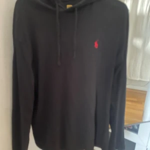 Svart hoodie från Polo Ralph Lauren - Svart hoodie från Polo Ralph Lauren med röd broderad logga på bröstet. Tröjan har huva med snörning och långa ärmar. Tillverkad i mjuk bomull som är skön att ha på sig. Perfekt för dig som gillar stilrena och klassiska plagg.