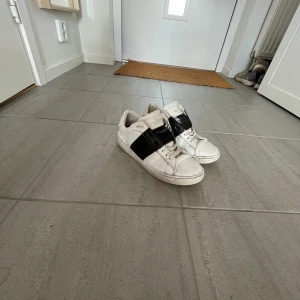 Valentino Garavani vita sneakers - Snygga vita sneakers från Valentino Garavani med en bred svart läderdetalj över mitten. Klassisk rund tå, platt sula och snörning framtill. Märkeslogga på plösen och stilren design som sticker ut. Perfekta för dig som gillar exklusiva streetwear-vibbar. Hål under båda sulorna men har täckt igen med två bra sular satt oavsett de regnar så läcker det inte in 👍🏻 