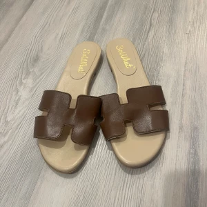 Bruna sandaler från SoWhat, stl 40 - Snygga bruna sandaler från SoWhat i storlek 40. De har en bred rem i brunt syntetmaterial med coolt utskuret mönster och en beige sula. Perfekta för varma dagar och enkel att matcha med allt.