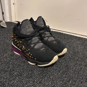 Nike LeBron 16 svart/guld sneakers - Snygga Nike LeBron 16 sneakers i svart med guldiga detaljer och lila inslag. Ovandelen har ett mönstrat tyg och tungorna pryds av ett lejonmotiv. Sulan är vit och lila med synlig dämpning. Perfekta för dig som gillar unika och sportiga skor.