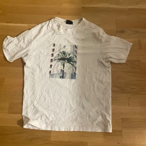 Vit t-shirt med palmtryck - Vit t-shirt med tryck av en palm och texten 'sit back and relax' på bröstet. Klassisk rund halsringning och korta ärmar. Perfekt för en chill och avslappnad stil. Materialet känns som bomull och trycket ger en cool vibe.