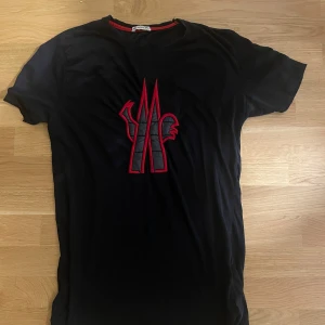 Svart Moncler t-shirt med röd logga - Svart t-shirt från Moncler i bomull med stor, upphöjd logga i svart och rött på bröstet. Klassisk rund halsringning med röd detalj på insidan av kragen. Perfekt för dig som gillar streetstyle och vill sticka ut med en fet logga.