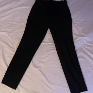 Svarta kostymbyxor med smal passform - Snygga svarta kostymbyxor med smal passform och raka ben. Klassisk design med fickor fram och knappar bak. Perfekta för en stilren look och passar till skjorta eller t-shirt. Materialet känns mjukt och följsamt.