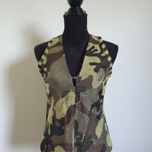 Camouflage topp från Jucca strl 36 - Snygg korsett-topp från Jucca i camouflage-mönster med guldiga inslag. Toppen är ärmlös, har djup v-ringning och dragkedja i ryggen. Materialet är glittrigt och ger en cool look. Perfekt för dig som vill sticka ut med en edgy stil. Nypris 2000 kr