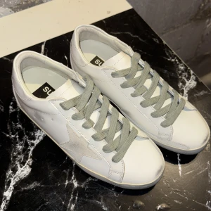 Golden Goose vita sneakers med stjärna - Golden Goose sneakers i vitt skinn med grå mockadetaljer och grå snörning. Klassisk låg modell med rund tå och platt sula. Sidan pryds av en ikonisk stjärna i mocka och baksidan har Golden Goose-logga. Perfekta för en clean och trendig look.UTFÖRSÄLJNING GÄLLER TILL 10 OKTOBER !!!
