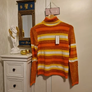 Randig polotröja från JIANGQIANQIAN - Snygg polotröja med breda ränder i orange, gul, vit och bruna toner från JIANGQIANQIAN. Tröjan har lång ärm, ribbad polokrage och är stickad i mjukt material. Perfekt för dig som gillar färg och retrovibbar. Köpt I Italien.