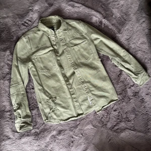 Ljusgrön jeansskjorta med slitningar - Snygg ljusgrön jeansskjorta från H&M med slitna detaljer och två bröstfickor. Skjortan har knappar framtill och långa ärmar. Perfekt för en avslappnad och trendig look.