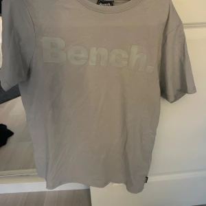Ljusgrå t-shirt från Bench i bomull - Snygg ljusgrå t-shirt från Bench med stor logga framtill. Klassisk passform och rund halsringning. Tillverkad i mjuk bomull, perfekt för en clean och enkel stil.