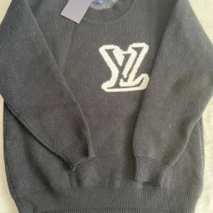 Svart stickad tröja Louis Vuitton - Svart stickad tröja från Louis Vuitton med stort vitt LV-logo på bröstet. Tröjan har rund halsringning, ribbade muddar och är långärmad. Perfekt för dig som vill ha en lyxig och stilren look med exklusiv känsla.