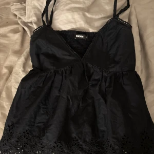 Svart ärmlös topp från BikBok - Supersöt topp från BikBok med smala axelband, v-ringning och broderade blommiga detaljer längst ner. Är gjort av ett tunt, skönt bomullstyg. Perfekt för varma dagar eller på kallare tider med en kofta, och enkelt att styla.🤍