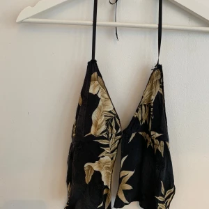 Svart blommig öppen rygg linnetopp - Snygg svart lätt linnetopp med tropiskt blommigt mönster i beige och guldtoner. Toppen har smala knytband i nacken och öppen rygg, vilket ger en cool och luftig känsla. Perfekt för varma dagar och chill vibes. Finns två olika sätt att styla den på med rosett i mak eller fram. Aldrig använd.