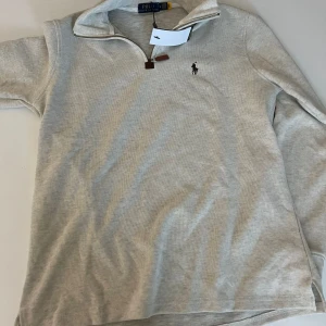Grå half zip tröja från Polo Ralph Lauren - Snygg ljusgrå långärmad tröja från Polo Ralph Lauren med klassisk broderad logga på bröstet. Tröjan har en half zip-dragkedja med läderdetalj och hög krage. Perfekt för lager-på-lager och chill stil.