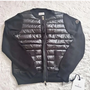 Svart quiltad cardigan jacka Moncler - Jag säljer min Moncler cardigan 🖤. Den är svart med quiltad, lite glansig front, dragkedja fram och två söta fickor med knappar. Stickade ärmar och ribbade muddar gör den både mysig och stilren. Självklart med den klassiska Moncler-loggan på ärmen 🤍. Den är perfekt om du vill ha en lyxig men ändå vardagssnygg look ✨. Säljer bara för att jag har lite för många jackor 🙈.